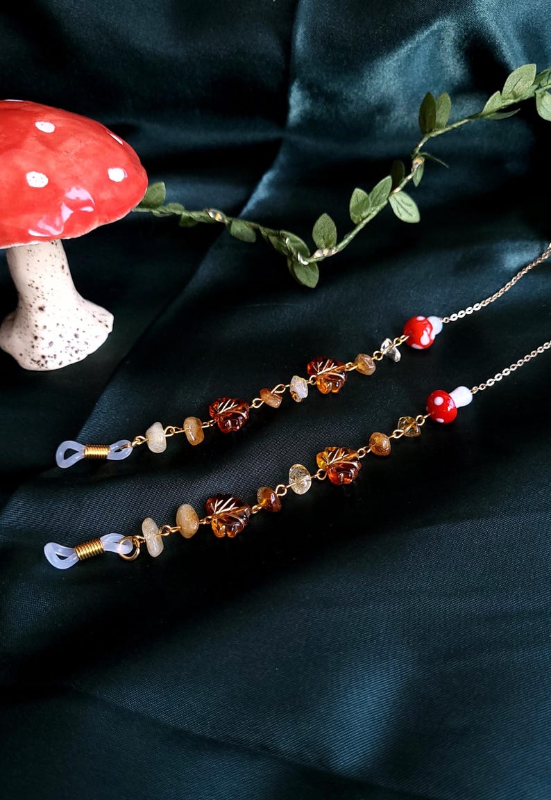 May include: Two eyeglass chains with gold-colored chains, amber-colored leaf charms, and small, round, red and white mushroom charms. The chains also feature small, light-colored stone beads. The background is a dark green fabric.