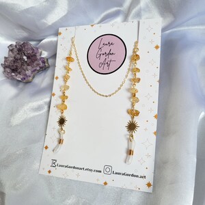 May include: Gold-toned eyeglass chain with citrine and quartz crystal accents. The chain features a sunburst charm and the text "Laura Gordon Art" on the product display card. The background is a white satin fabric.