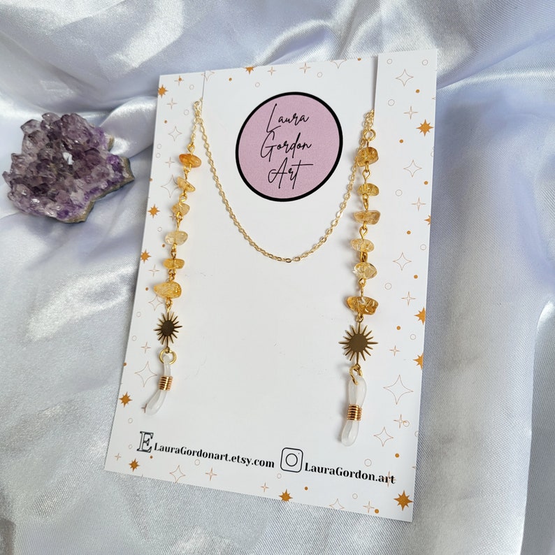 May include: Gold-toned eyeglass chain with citrine and quartz crystal accents. The chain features a sunburst charm and the text "Laura Gordon Art" on the product display card. The background is a white satin fabric.