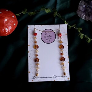 May include: A gold-colored chain necklace and earrings set. The earrings feature a series of amber-colored leaf charms and gemstones. The necklace and earrings are displayed on a white card with the text "Laura Gordon Art". A red mushroom is visible in the background.
