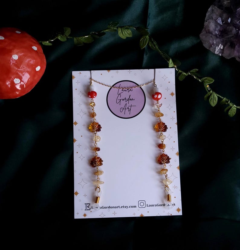 May include: A gold-colored chain necklace and earrings set. The earrings feature a series of amber-colored leaf charms and gemstones. The necklace and earrings are displayed on a white card with the text "Laura Gordon Art". A red mushroom is visible in the background.