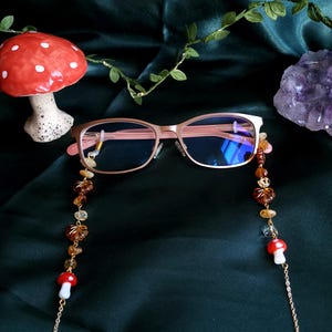 May include: Rose gold eyeglasses with a beaded chain featuring amber-colored beads and mushroom charms. The glasses have a rectangular frame and are set against a dark green background, with a ceramic mushroom and a crystal visible.