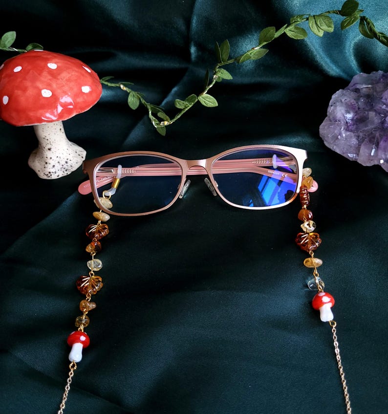 May include: Rose gold eyeglasses with a beaded chain featuring amber-colored beads and mushroom charms. The glasses have a rectangular frame and are set against a dark green background, with a ceramic mushroom and a crystal visible.