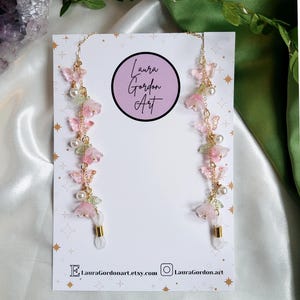 May include: Eyeglass chains featuring pink flower and butterfly details, white pearls, and gold-coloured chains. The chains are displayed on a white card with the text "Laura Gordon Art" and social media details.