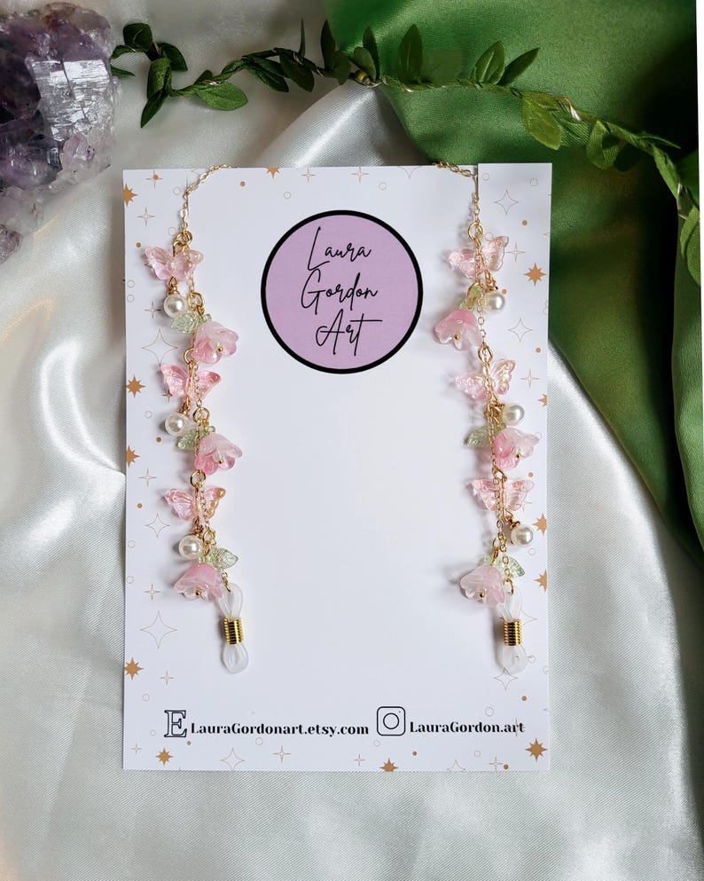 May include: Eyeglass chains featuring pink flower and butterfly details, white pearls, and gold-coloured chains. The chains are displayed on a white card with the text "Laura Gordon Art" and social media details.