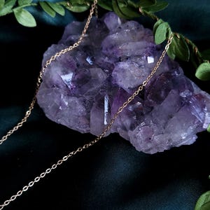 May include: A delicate gold chain necklace draped over a large, rough-cut amethyst crystal. The crystal is a deep purple color, and the necklace has a simple, elegant design. Green leaves are arranged around the crystal, creating a natural aesthetic.