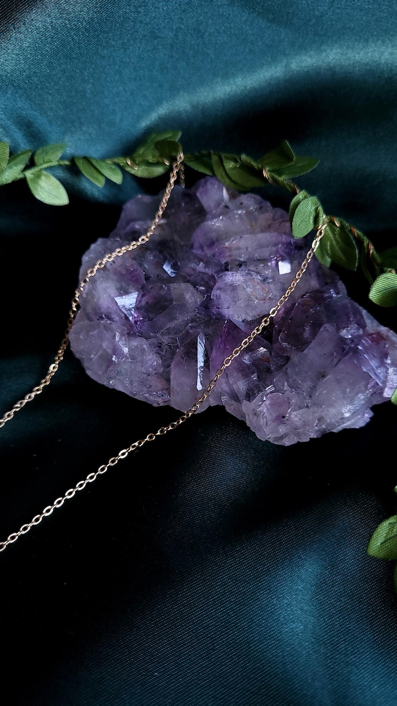May include: A delicate gold chain necklace draped over a large, rough-cut amethyst crystal. The crystal is a deep purple color, and the necklace has a simple, elegant design. Green leaves are arranged around the crystal, creating a natural aesthetic.