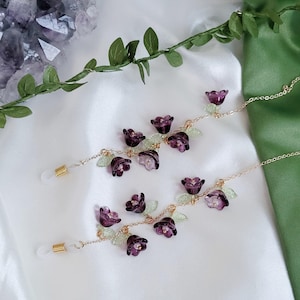 以下が含まれることがあります： ゴールドカラーのチェーンと紫色の花と緑の葉のアクセントが付いた2つのメガネチェーン。チェーンは、小さな半透明の紫色の花と明るい緑の葉で飾られています。メガネホルダーは白です。