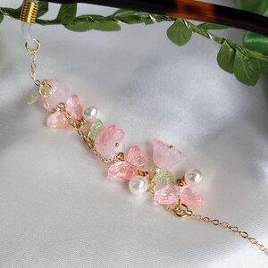 May include: An elegant eyeglass chain with a gold-tone chain and floral details. The chain features pink and green translucent flowers, faux pearls, and a gold-tone connector. The chain is approximately 25 cm long.