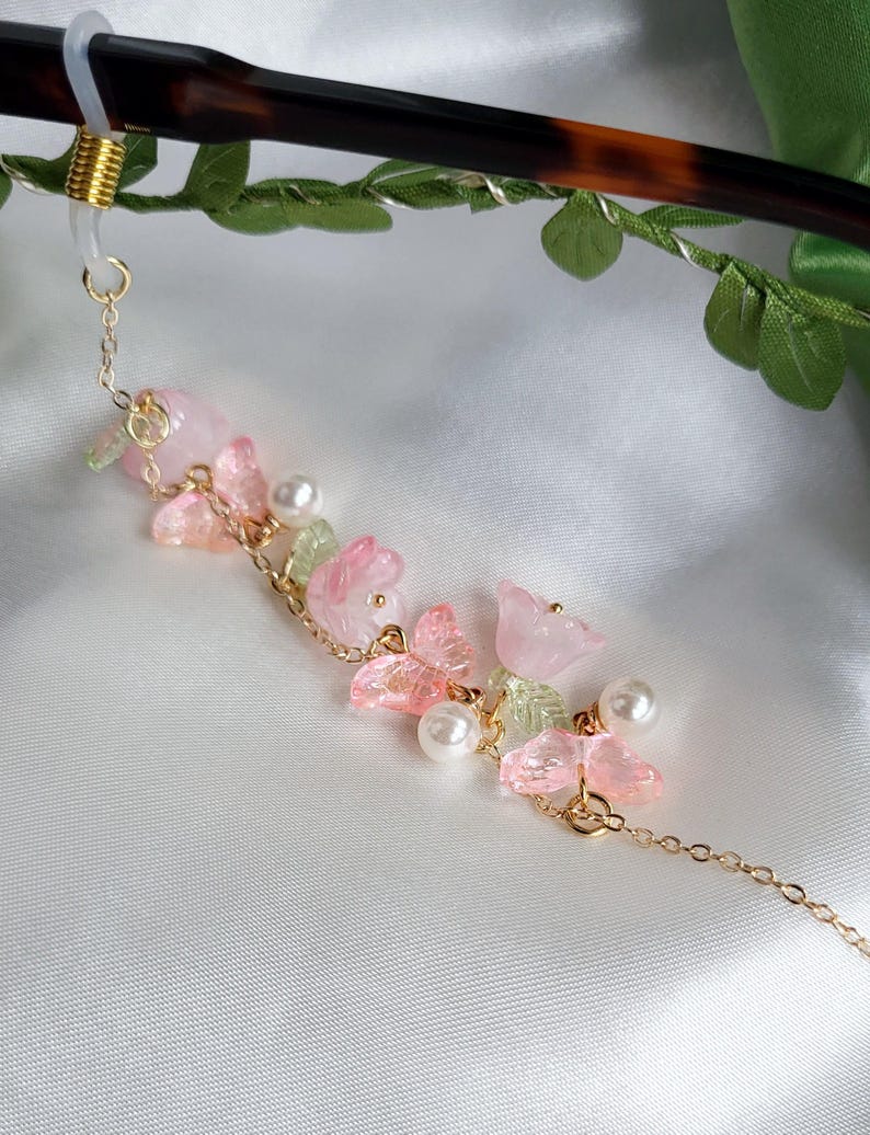 May include: An elegant eyeglass chain with a gold-tone chain and floral details. The chain features pink and green translucent flowers, faux pearls, and a gold-tone connector. The chain is approximately 25 cm long.