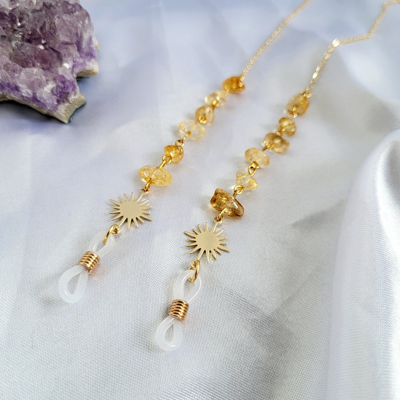 May include: Two gold-colored eyeglass chains with citrine gemstone beads and sunburst charms. The chains have white rubber loops for attaching to glasses. The background is a white, silky fabric.
