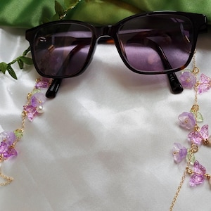 May include: Sunglasses with dark lenses are accessorised with a decorative chain. The chain features purple flowers, butterfly charms, green accents, and faux pearls, creating an elegant accessory.