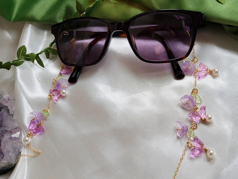 May include: Sunglasses with dark lenses are accessorised with a decorative chain. The chain features purple flowers, butterfly charms, green accents, and faux pearls, creating an elegant accessory.
