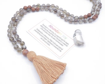May include: A brown and gray mala necklace with a tassel. The necklace is made of citrine quartz and sandalwood beads. The card reads "Abundant Joy" and includes information about the stones.