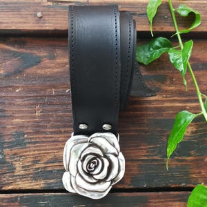 May include: A black leather belt featuring a large, three-dimensional silver-toned metal buckle shaped like a blooming rose. The belt strap is smooth with visible stitching along the edges and is secured to the buckle with two silver metal rivets.