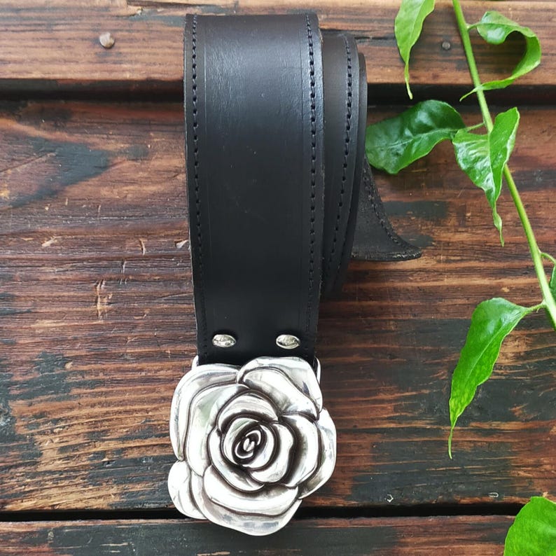May include: A black leather belt featuring a large, three-dimensional silver-toned metal buckle shaped like a blooming rose. The belt strap is smooth with visible stitching along the edges and is secured to the buckle with two silver metal rivets.