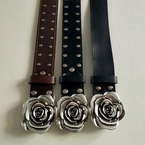 May include: Three belts arranged vertically, featuring a large, silver-toned metal rose-shaped buckle. The left belt is brown with two rows of silver studs, while the center belt is black with two rows of studs and the right belt is plain black.