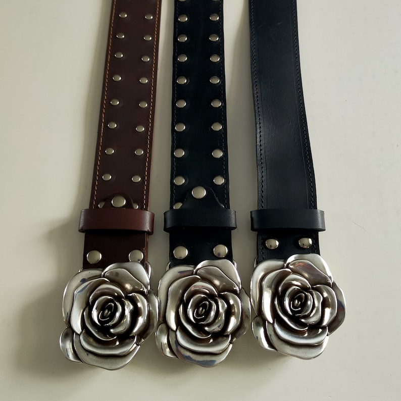 May include: Three belts arranged vertically, featuring a large, silver-toned metal rose-shaped buckle. The left belt is brown with two rows of silver studs, while the center belt is black with two rows of studs and the right belt is plain black.