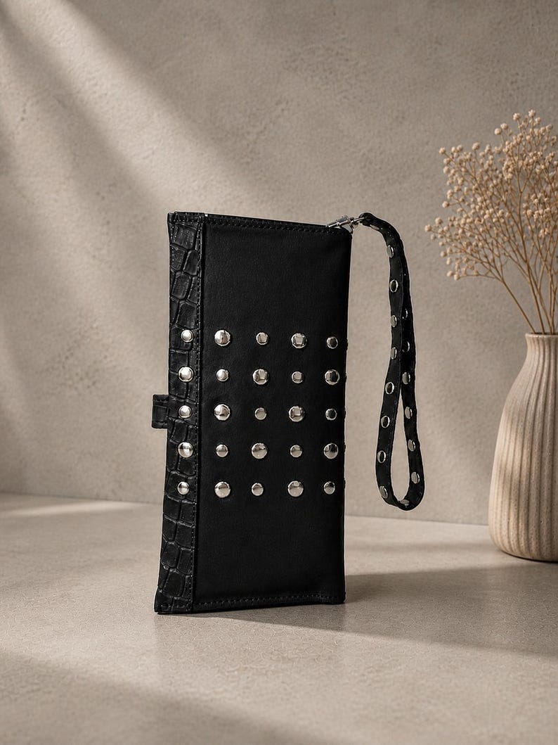 May include: A black rectangular clutch wallet featuring a vertical panel with a textured crocodile-embossed pattern and a main section adorned with a grid of silver-toned metal studs. The wallet includes a matching wrist strap with silver studs.