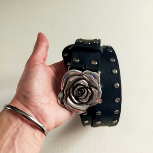May include: A black leather belt featuring a large, three-dimensional silver-toned metal rose buckle. The belt strap is adorned with evenly spaced, circular silver-toned metal studs along its length, providing a consistent metallic accent.