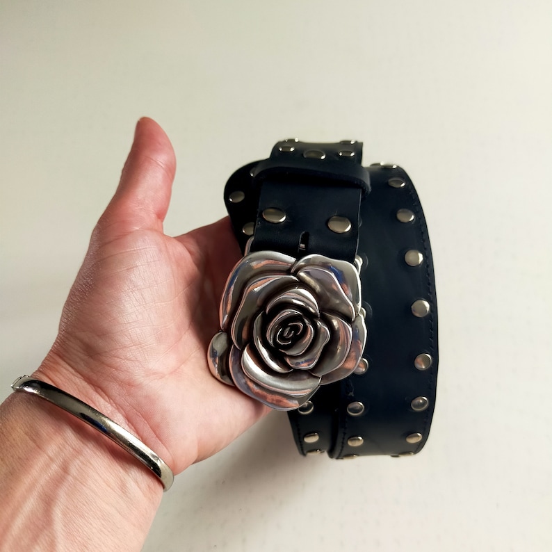 May include: A black leather belt featuring a large, three-dimensional silver-toned metal rose buckle. The belt strap is adorned with evenly spaced, circular silver-toned metal studs along its length, providing a consistent metallic accent.