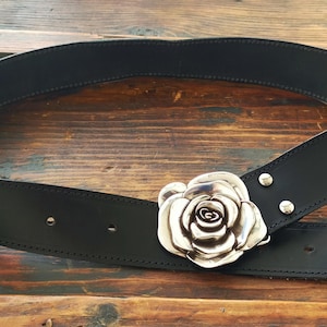 May include: A black leather belt featuring a large, three-dimensional silver-toned metal rose buckle. The belt strap has visible stitching along the edges and two silver-toned metal rivets securing the rose buckle to the leather material.