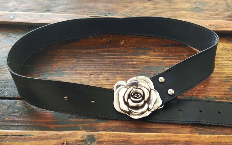 May include: A black leather belt featuring a large, three-dimensional silver-toned metal rose buckle. The belt strap has visible stitching along the edges and two silver-toned metal rivets securing the rose buckle to the leather material.