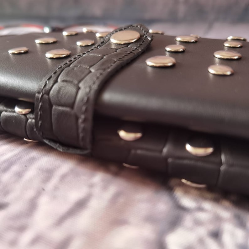 May include: A dark brown wallet featuring a textured crocodile-patterned strap closure and numerous polished silver-tone metal studs embedded across the surface. The wallet is constructed with a smooth finish and includes a central snap button.
