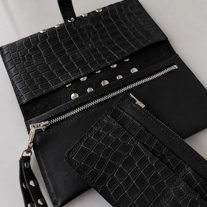 May include: A black clutch wallet with a crocodile-embossed texture, featuring a snap-button flap closure, silver-tone metal studs, a zippered compartment, and a detachable wrist strap with matching silver studs.