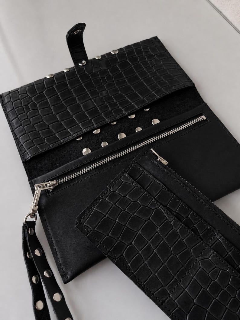 May include: A black clutch wallet with a crocodile-embossed texture, featuring a snap-button flap closure, silver-tone metal studs, a zippered compartment, and a detachable wrist strap with matching silver studs.