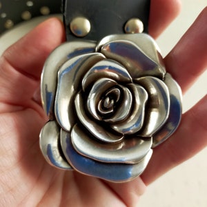 May include: A black belt featuring a large, three-dimensional silver-toned metal rose buckle. The rose is constructed with layered, curved petals and is attached to the black strap with two visible silver-toned metal rivets at the top.