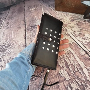 May include: A black rectangular wallet featuring a series of silver-toned metal studs across the front surface. The wallet includes a dark brown textured strap with a snap button closure and a thin black leather pull tab attached to the zipper.