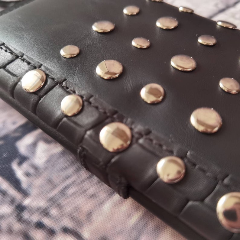 May include: A black wallet featuring a smooth leather surface adorned with numerous polished silver-tone metal studs. The edge of the wallet displays a textured, embossed pattern resembling crocodile skin, complemented by visible stitching.