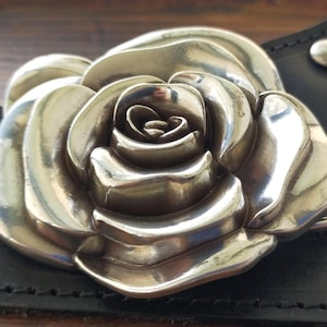 May include: A large, polished silver-toned metal belt buckle shaped like a blooming rose with layered petals. The buckle is attached to a black leather belt strap featuring visible stitching and a small silver-toned metal rivet near the edge.