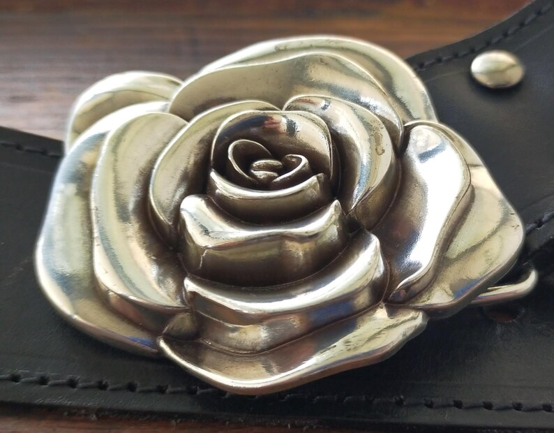 May include: A large, polished silver-toned metal belt buckle shaped like a blooming rose with layered petals. The buckle is attached to a black leather belt strap featuring visible stitching and a small silver-toned metal rivet near the edge.
