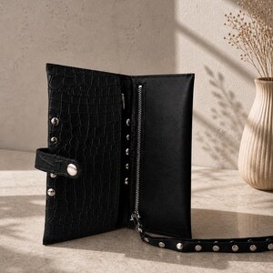 May include: A black wallet with a crocodile-embossed texture, featuring a silver-toned zip compartment, a snap-button closure, and a matching wrist strap adorned with silver metal studs along the exterior and the strap edge.