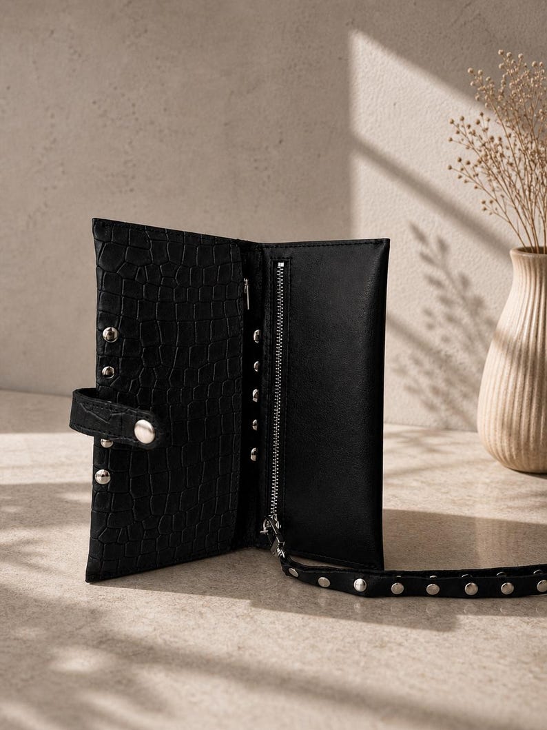 May include: A black wallet with a crocodile-embossed texture, featuring a silver-toned zip compartment, a snap-button closure, and a matching wrist strap adorned with silver metal studs along the exterior and the strap edge.