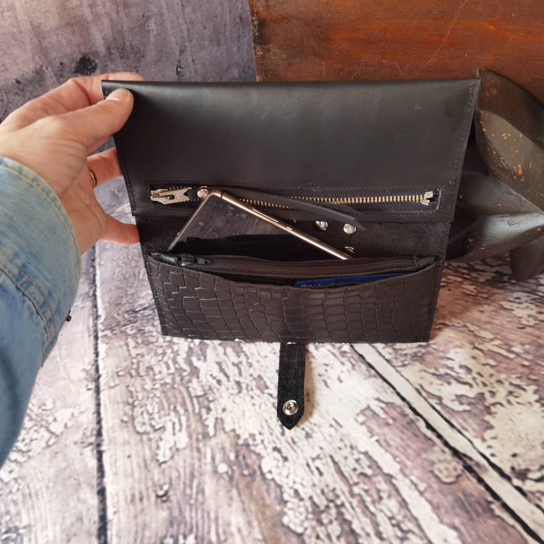May include: A black leather wallet featuring a flap closure with a snap button, a textured crocodile-embossed front panel, and an interior compartment containing a smartphone and a zippered pocket with a silver-toned metal zipper pull.