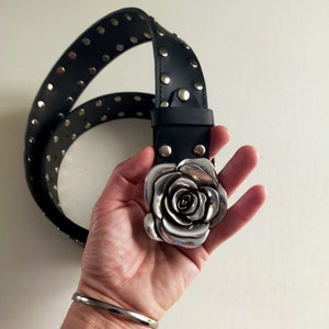 May include: A black leather belt featuring a large, three-dimensional silver-toned metal rose buckle. The strap is adorned with a single row of evenly spaced, circular silver-toned metal studs that run along the length of the belt.