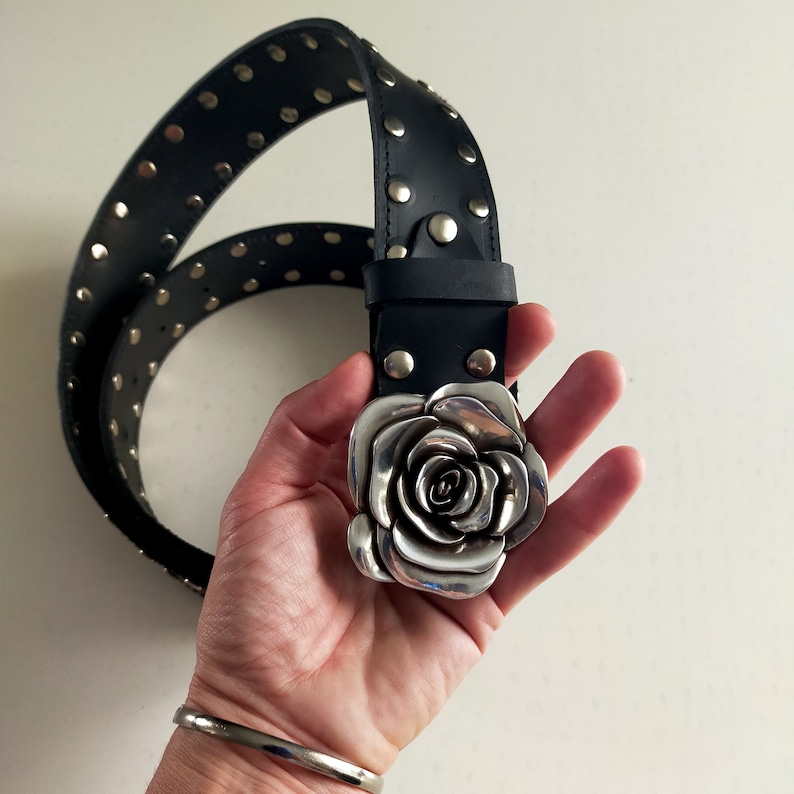 May include: A black leather belt featuring a large, three-dimensional silver-toned metal rose buckle. The strap is adorned with a single row of evenly spaced, circular silver-toned metal studs that run along the length of the belt.