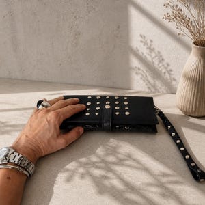 May include: A black rectangular clutch wallet featuring a textured crocodile-embossed finish and silver-toned metal studs arranged in rows across the front flap. The wallet includes a matching black wrist strap adorned with silver studs.