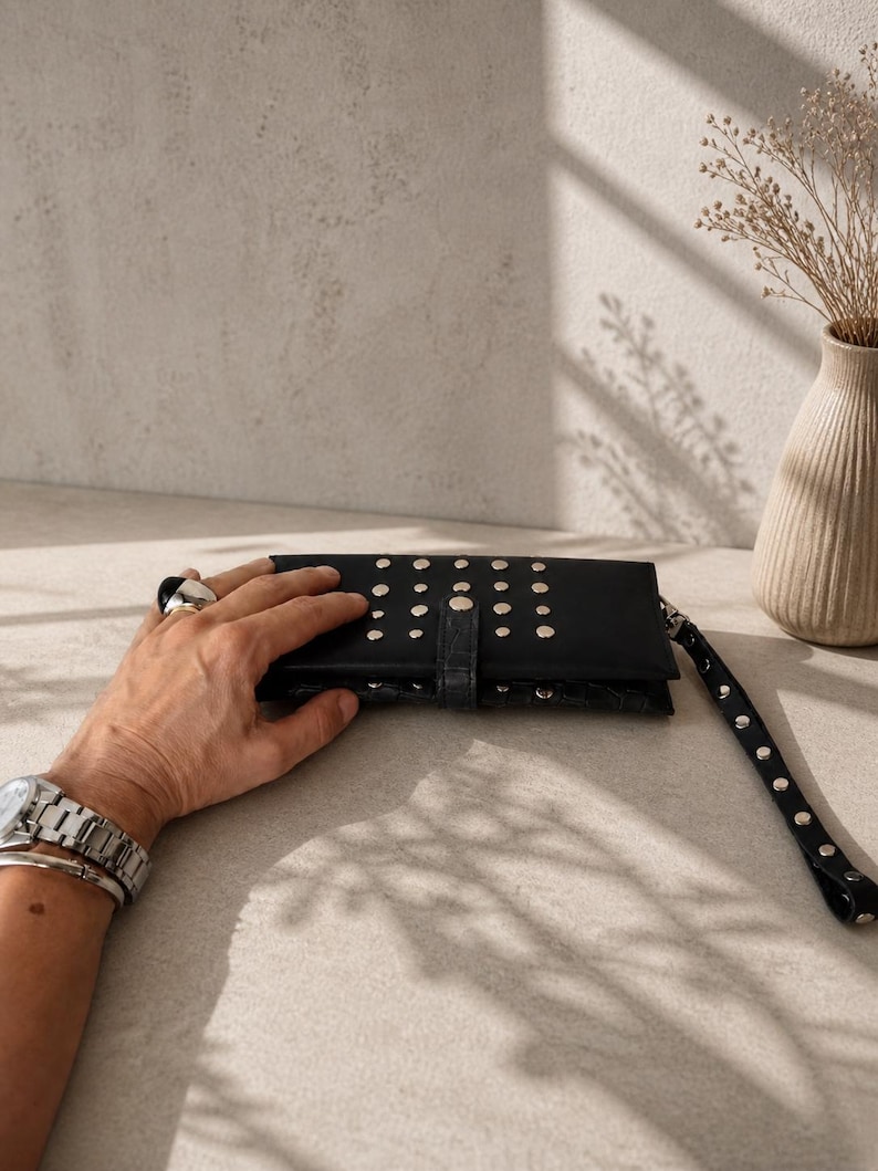 May include: A black rectangular clutch wallet featuring a textured crocodile-embossed finish and silver-toned metal studs arranged in rows across the front flap. The wallet includes a matching black wrist strap adorned with silver studs.