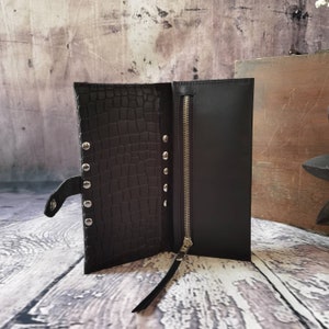 May include: A black wallet featuring a textured crocodile-embossed pattern on one side and a smooth finish on the other. The wallet includes a vertical zippered compartment with a metallic zip and a row of silver-toned studs along the centre fold.