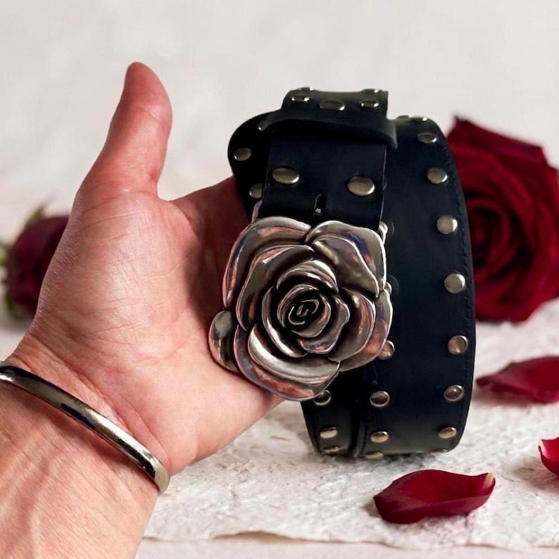 May include: A black leather belt featuring a large, three-dimensional silver-toned metal rose buckle. The belt strap is adorned with a series of evenly spaced, circular silver-toned metal studs running along the length of the band.