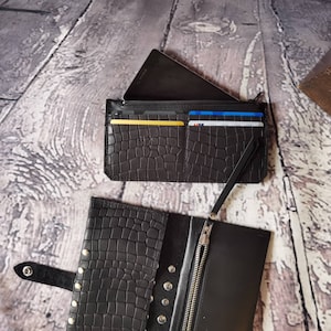May include: Two black wallets with a crocodile-embossed texture. One wallet is open, showing a silver zip, a snap button closure, and multiple metal studs. The other wallet is closed, featuring card slots holding several credit cards.