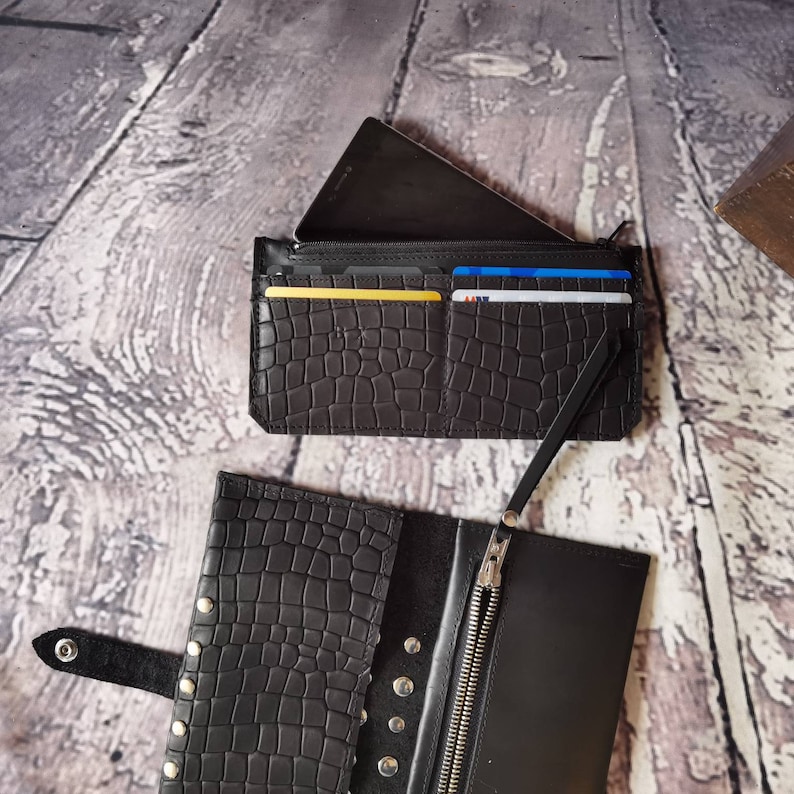 May include: Two black wallets with a crocodile-embossed texture. One wallet is open, showing a silver zip, a snap button closure, and multiple metal studs. The other wallet is closed, featuring card slots holding several credit cards.