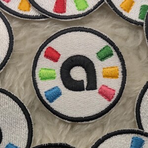 May include: Circular embroidered patches with a white base, black border, and a central black letter "a". The patches have colourful rectangular accents in red, green, blue, yellow, and pink. The patches are arranged on a fluffy, light-coloured surface.