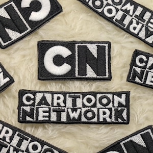 Może przedstawiać: Czarno-białe, haftowane naszywki z logo i tekstem Cartoon Network. Prostokątne naszywki mają czarne tło z białymi przeszyciami. Naszywki są rozrzucone na puszystej, kremowej powierzchni.