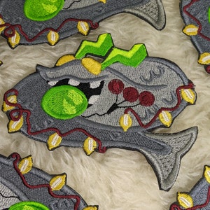 May include: Embroidered gray fish patch with a large green eye, open mouth, and a green lightning bolt on top. The fish is decorated with red berries and a string of yellow and white lights. The patches are on a white, fluffy surface.