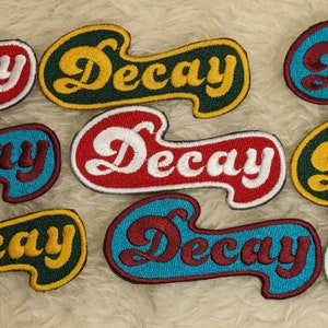 Può includere: Nove toppe ricamate con la parola "Decay" in un font retrò. Le toppe sono di vari colori, tra cui rosso, blu, verde e giallo.